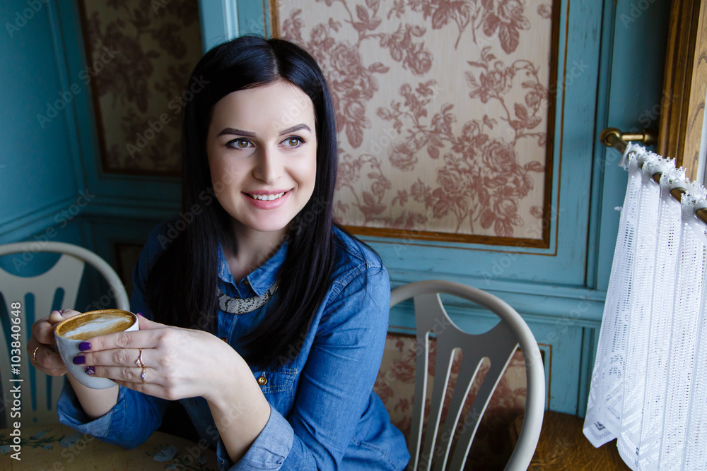 Beautiful happy young woman dressed in denim shirt sitting in the cafe drinking coffee and looking trough the window Beautiful happy young woman dressed in denim shirt sitting in the cafe drinking coffee and looking trough the window