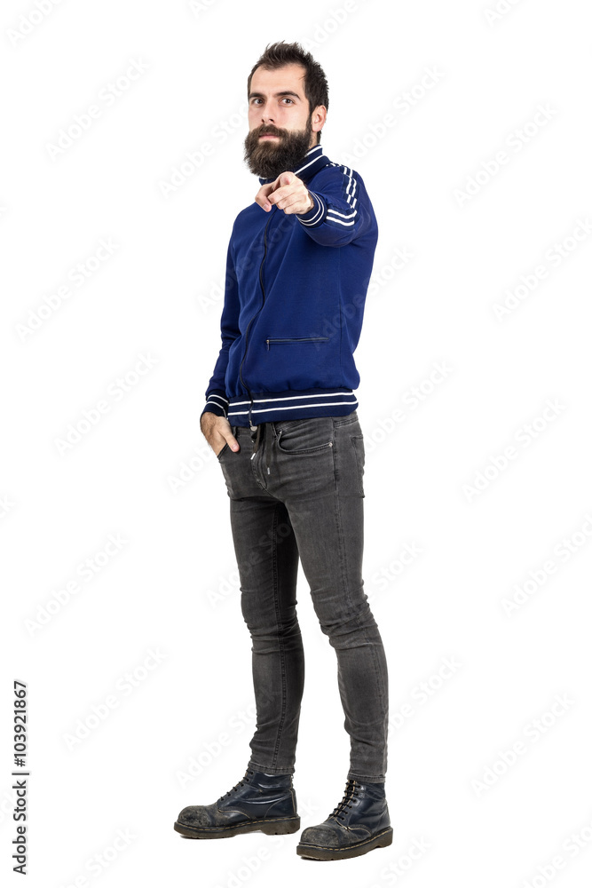 Confident serious hipster in blue tracksuit jacket pointing finger at camera. Full body length portrait isolated over white studio background. Confident serious hipster in blue tracksuit jacket pointing finger at camera. Full body length portrait isolated over white studio background.
