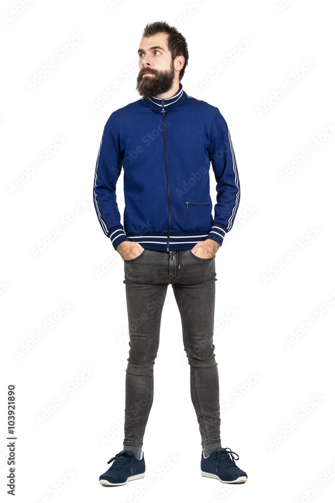 Serious bearded hipster in tracksuit jacket with hands in pockets looking away. Full body length portrait isolated over white studio background. Serious bearded hipster in tracksuit jacket with hands in pockets looking away. Full body length portrait isolated over white studio background.