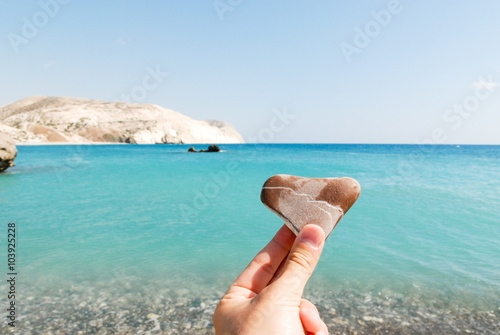 Hand holding a heart-shaped stone and Aphrodite bay in the background