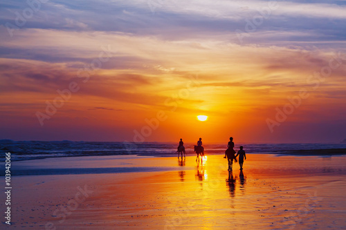 Black silhouette of happy family having horse riding adventure on sand sea beach on background of sunset sky. Active parents and people outdoor activity on tropical summer vacations with children. © Tropical studio