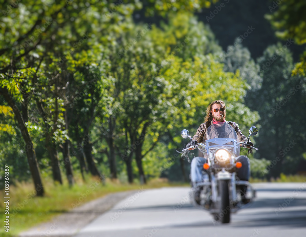 Biker with long hair and beard in sunglasses and leather jacket on the road. Tilt shift lens blur effect