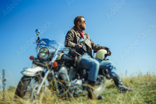 Portrait of a young man with beard sitting on his cruiser motorcycle and relaxing after driving. Man is wearing leather jacket and blue jeans. Low point of view. Tilt shift lens blur effect