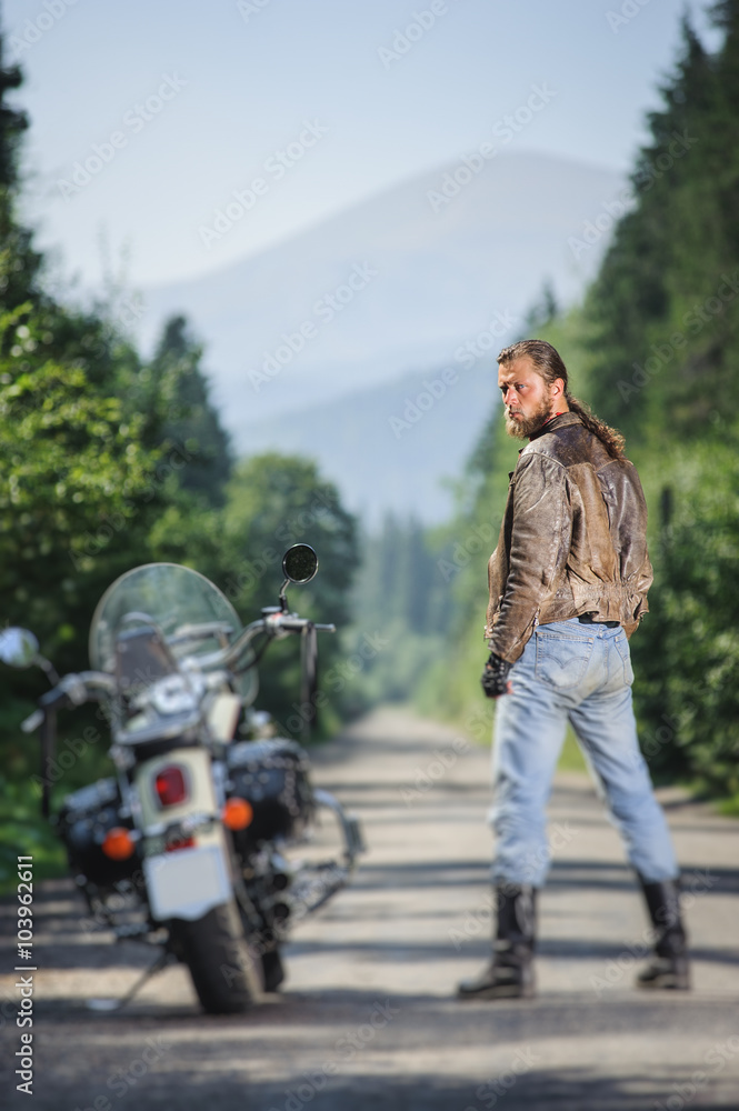 Biker with long hair wearing leather jacket blue jeans boots and gloves  standing near his cruiser