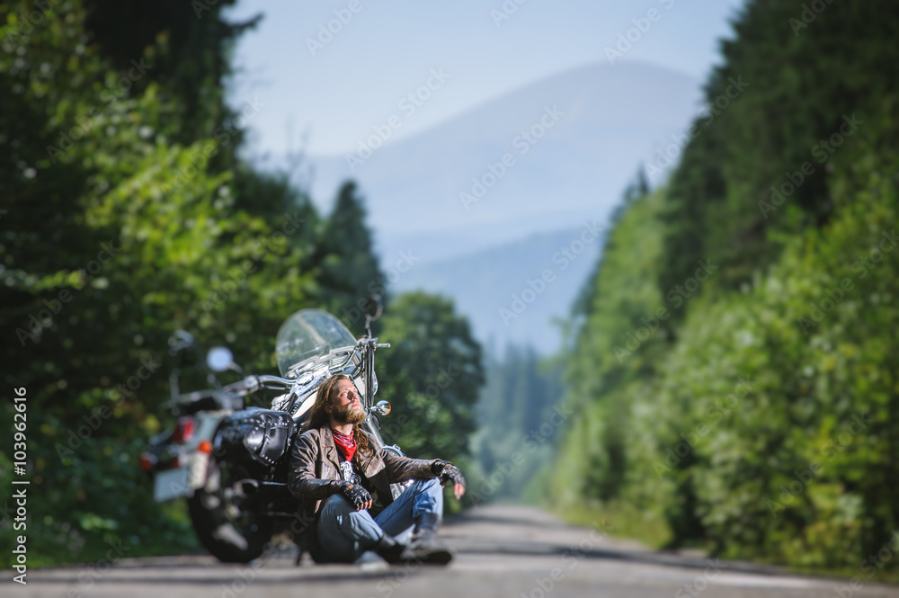 Handsome biker with beard and long hair sitting next to a traveler motorcycle on an open road looking to the sun. Guy is wearing leather jacket and blue jeans. Sunny summer day. Tilt shift soft effect Handsome biker with beard and long hair sitting next to a traveler motorcycle on an open road looking to the sun. Guy is wearing leather jacket and blue jeans. Sunny summer day. Tilt shift soft effect