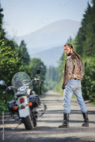 Biker with long hair wearing leather jacket blue jeans boots and gloves standing near his cruiser motorcycle on the open road. Looking to the camera. View from the back. Tilt shift soft effect