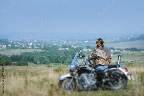 Brutal biker with beard wearing leather jacket and sunglasses sitting on his motorcycle on a sunny day, holding helmet. Horizontal picture. View from the back. Tilt shift lens blur effect