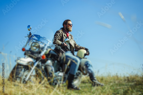 Portrait of a young happy biker with beard sitting on his cruiser motorcycle. Man is wearing leather jacket and blue jeans. Low point of view. Tilt shift lens blur effect