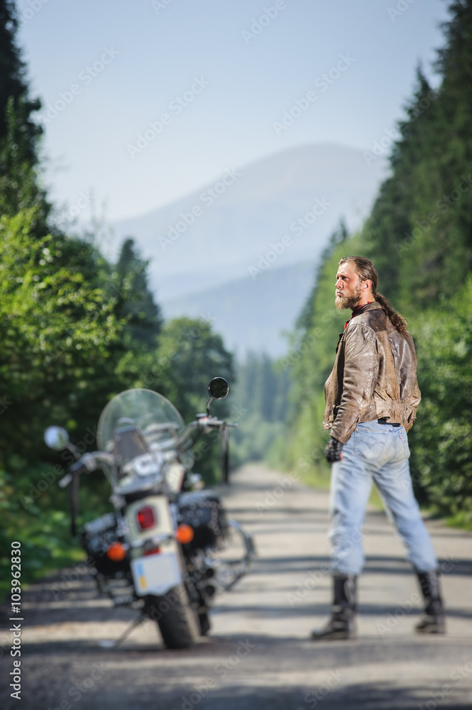 Handsome biker with long hair wearing leather jacket blue jeans leather boots and gloves standing near his custom made cruiser motorcycle on the open road. Looking to the sun. Tilt shift soft effect