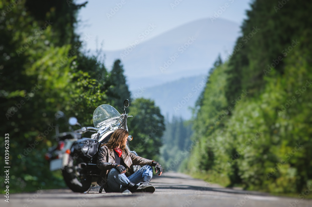 Handsome biker with beard and long hair sitting next to his custom made cruiser motorcycle on an open road. Guy is wearing leather jacket and blue jeans, looking into distance. tilt shift soft effect