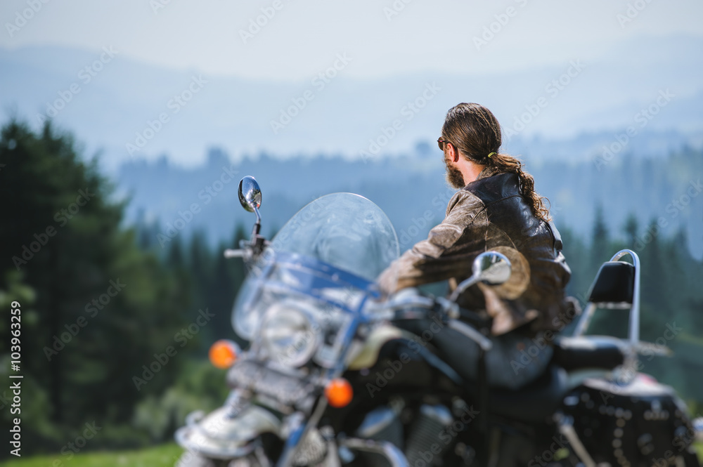 Portrait of long-haired guy in sunglasses jeans and a leather jacket sitting on a black custom motorcycle and looking into distance. Sunny day in the mountains. View from the back. Tilt blur effect