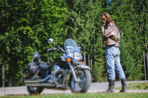 Handsome biker with long hair and beard standing near his custom made cruiser motorcycle looking down. Biker is wearing leather jacket and sunglasses on sunny day. tilt shift soft effect