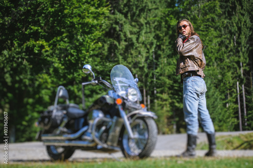 Handsome man with long hair and beard standing near his custom made cruiser motorcycle and looking to the sun. Biker is wearing leather jacket and sunglasses on sunny day. tilt shift soft effect