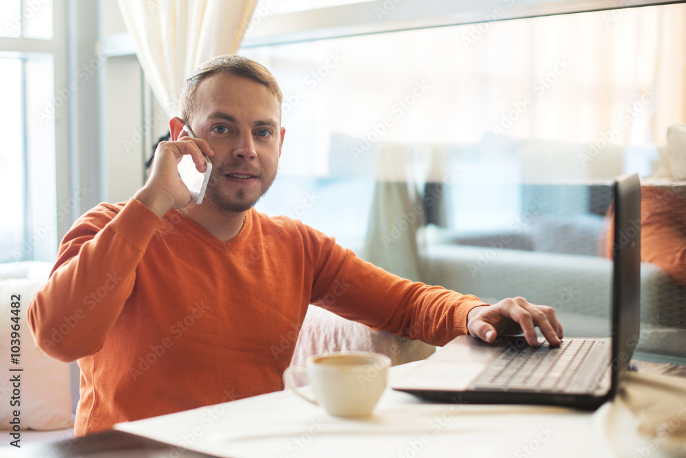 Handsome young man working with notebook, talking on the phone, looking at camera, while enjoying coffee in cafe