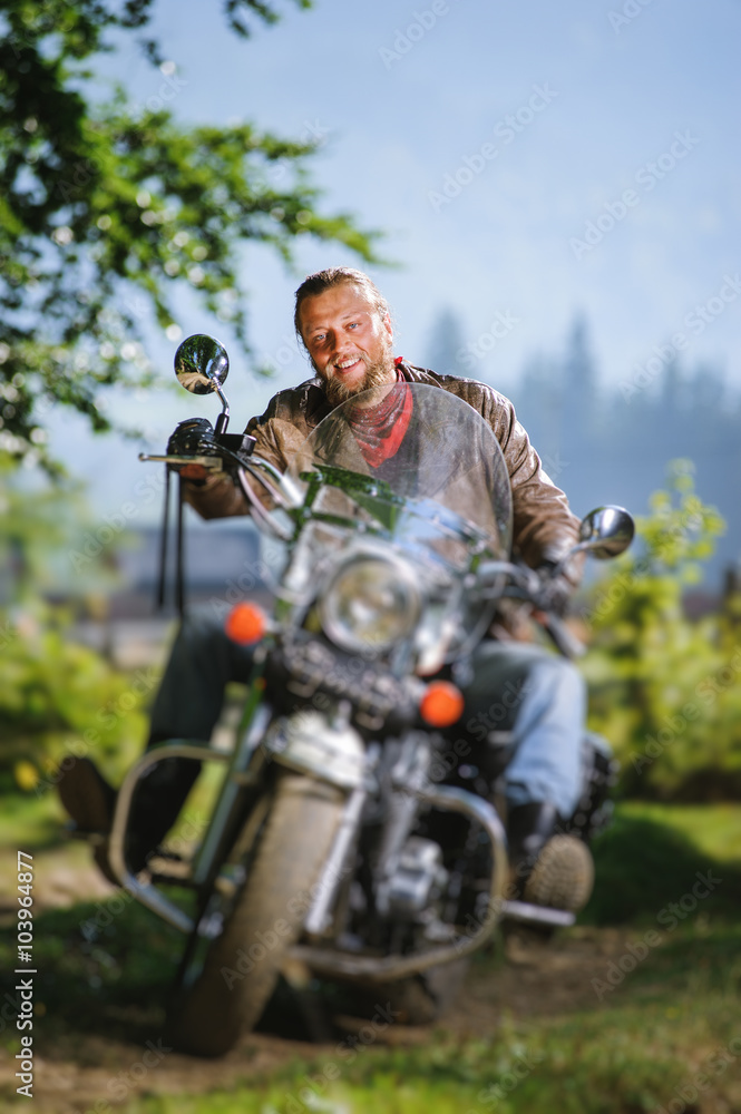 young biker with beard driving his cruiser motorcycle in the forest and smiling. Man is wearing leather jacket and blue jeans.  Tilt shift lens blur effect