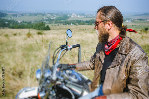 Portrait of biker with long hair and beard in a leather jacket and sunglasses sitting on his bike on the grassy field. Looking into the distance. Side view. Tilt shift lens blur effect