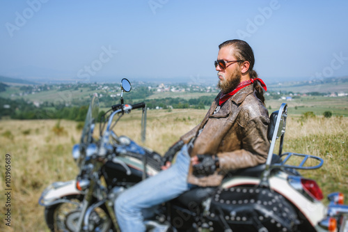 Portrait of biker with long hair and beard in a leather jacket and sunglasses sitting on his bike and relaxing on the grassy field. Looking into the distance. Side view. Tilt shift lens blur effect © anatoliy_gleb