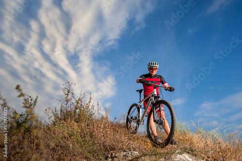 Fototapeta Naklejka Na Ścianę i Meble -  Mountain Bike cyclist resting near his bike in nature on sunny day. Low angle view with cloudy sky on the background. Cross country biking.