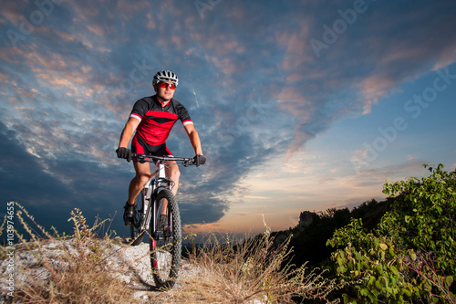 Happy man on a mountain bike races downhill in the nature against blue cloudy evening sky. Cyclist is wearing red sportswear helmet gloves and red glasses. Cross country biking.
