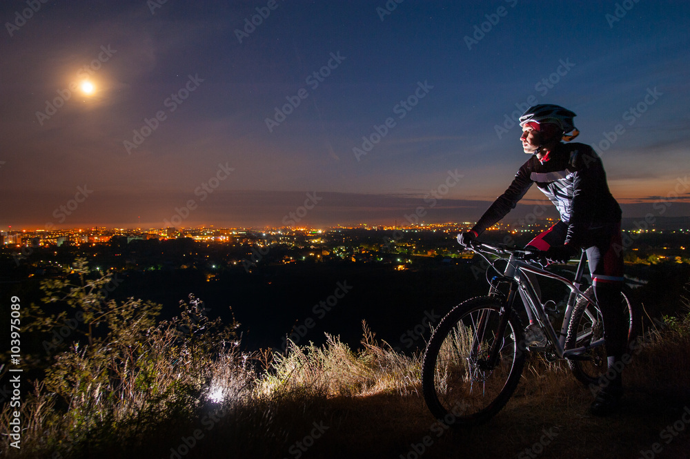 Young cyclist with mountain bike on top of the hill observing the night city view and full moon in the sky