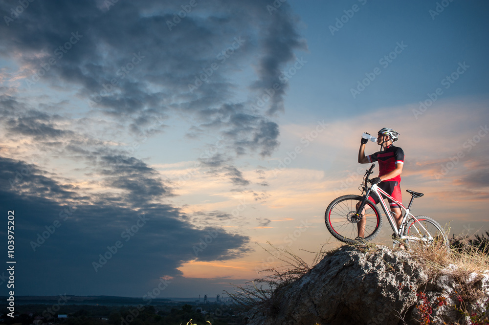 Obraz premium Cross country biker drinking water on top of a mountain with bike in the evening, sky background. side view