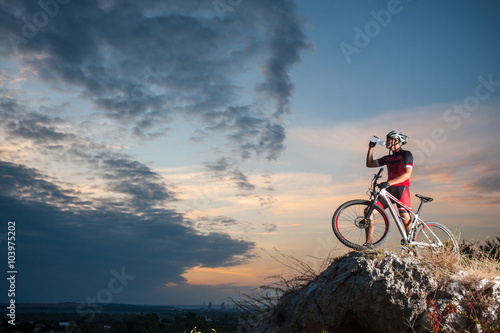 Cross country biker drinking water on top of a mountain with bike in the evening, sky background. side view