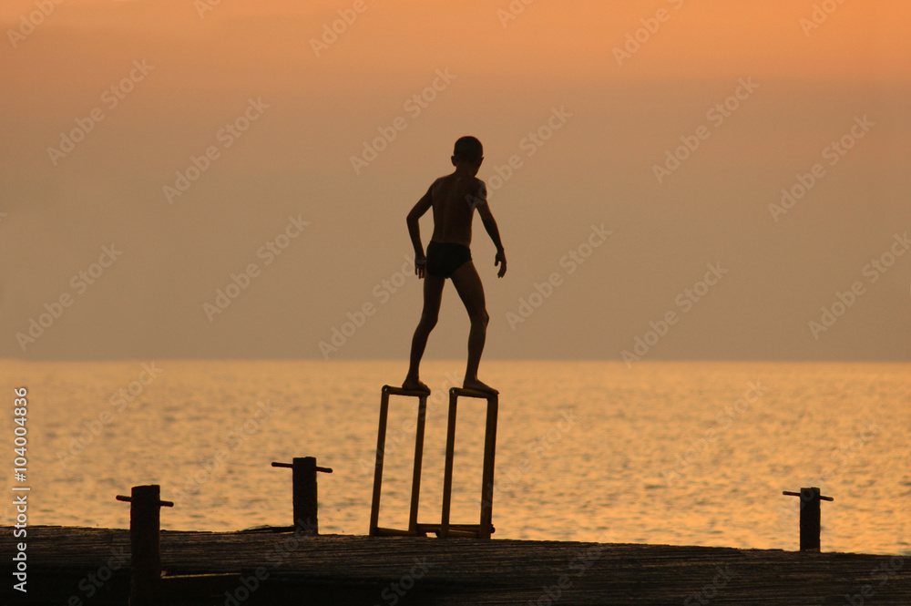 Fototapeta premium silouette of a boy ready to dive from pierce in front of brightl
