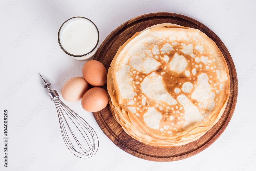 Thin pancakes on a round wooden stand with a whisk, and ingredients on a white background Thin pancakes on a round wooden stand with a whisk, and ingredients on a white background