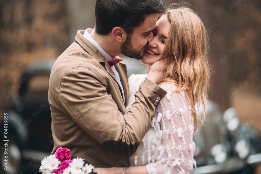 Romantic fairytale wedding couple kissing and embracing in pine forest near retro car.