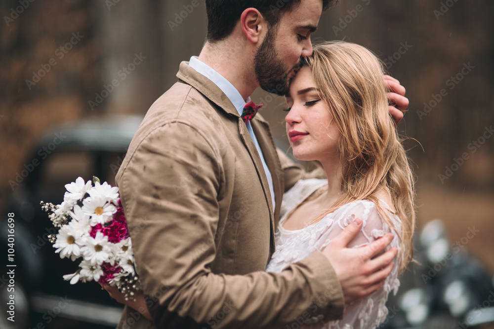 Fototapeta premium Romantic fairytale wedding couple kissing and embracing in pine forest near retro car.
