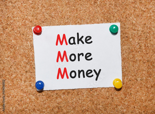 The phrase Make More Money typed on a white piece of note paper and pinned to a cork notice board