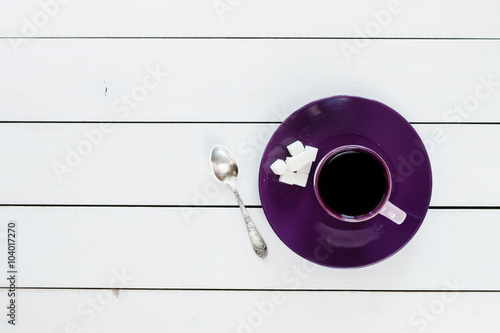 cup of tea or coffee on violet plate, silver tea spoon, lump sugar on white colored wooden table, top view