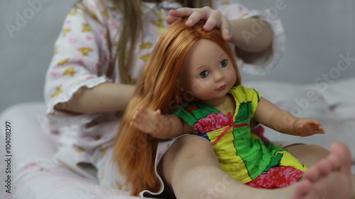 Girl playing with doll sitting on the bed, she braids her hair
