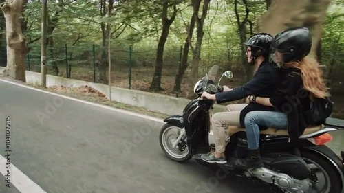 Slow motion side shot (60 fps) of young Caucasian couple riding their scooter on beautiful country road through forest