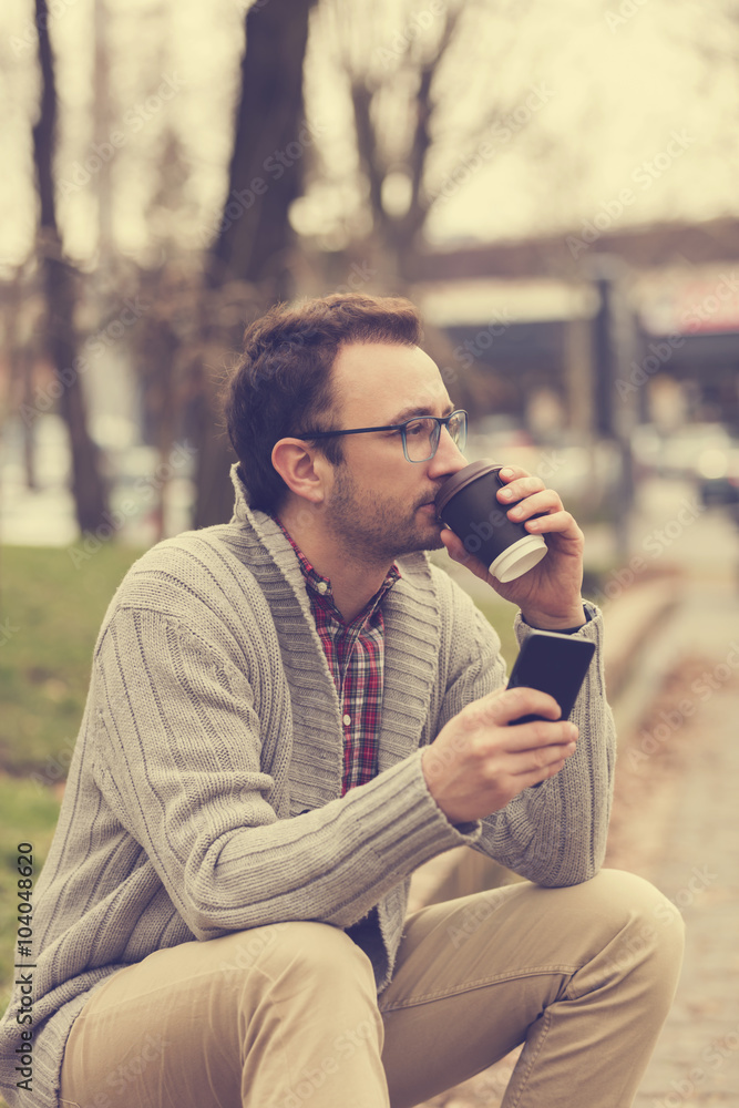 Hipster man sitting outdoors and drinking coffee while using his cellphone. Hipster man sitting outdoors and drinking coffee while using his cellphone.