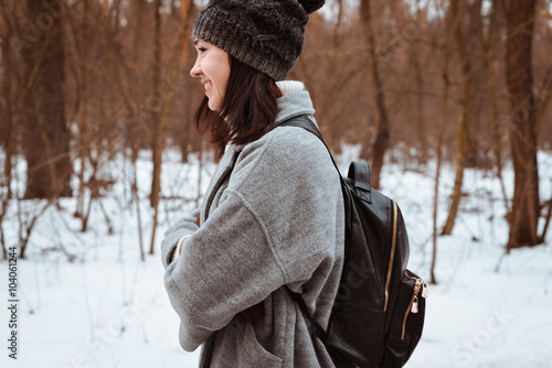 Portrait of a happy beautiful girl with brown hair in  winter forest.Attractive brunette woman dressed in a hipster style with black stylish leather backpack wearing grey coat and white pullover.