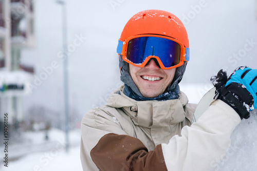 Man snowboarder wearing orange helmet, grey jacket, black and blue gloves standing with snowboard © Sergey