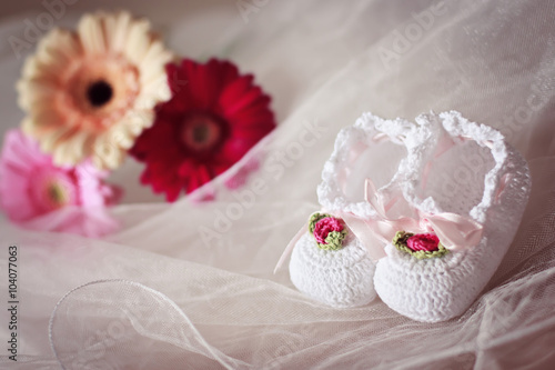 Fototapeta Naklejka Na Ścianę i Meble -  White knit baby booties decorated with flowers and ribbons  with gerberas in the background