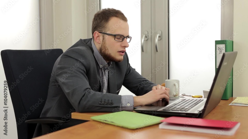 Young, handsome men sitting behind the desk and in the same time he is using computer. He is bored because of his work and he is getting sleep Young, handsome men sitting behind the desk and in the same time he is using computer. He is bored because of his work and he is getting sleep