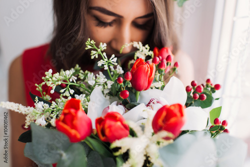 beautiful young girl in a red dress at a window with a wreath on his head and a bouquet of flowers, enjoying the smell of flowers