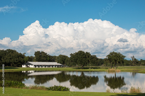 White equine horse barn on farm ranch with reflection in water pond lake and blue sky and white puffy idyllic clouds