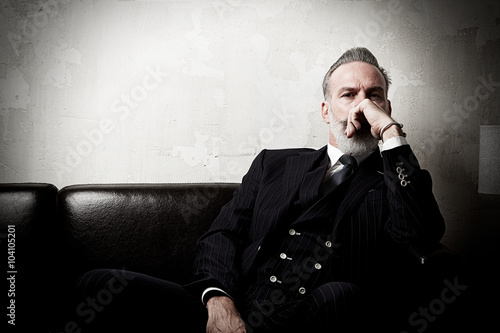 Foto Portrait of adult gentleman wearing trendy suit and sitting modern studio on leather sofa against the empty concrete wall