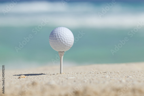 Golf ball on tee in sand beach with ocean waves behind
