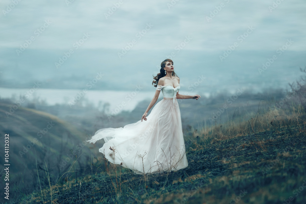Fantasy girl in white beige vintage dress long flying pastel train stands on top of mountain. Woman Shabby chic boho style.  fashion model, creative gown silk fabric flutter in wind. Cloudy autumn sky