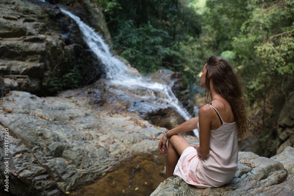 The beautiful fashionable girl with long hair, sits on surface stones, dressed in a light white dress, finding on tropical falls of the island Samui. Sunny day and happy smile.