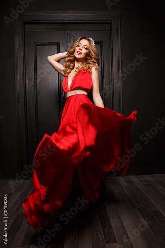 The queen of the ball with diadem on her head and gorgeous body is very passionate and stunning in the elegant red evening fluttering dress with deep neckline