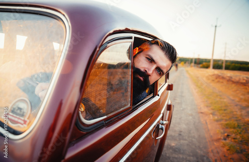 brutal bearded man with a mustache in a shirt, pants with suspenders with a girl with dark hair and big lips with bright red lipstick in a short dress and heels near retro car at sunset