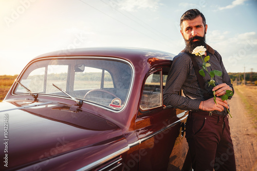 brutal bearded man with a mustache in a shirt, pants with suspenders stands leaning on the hood of a retro car holding a white rose and looking at the sunset