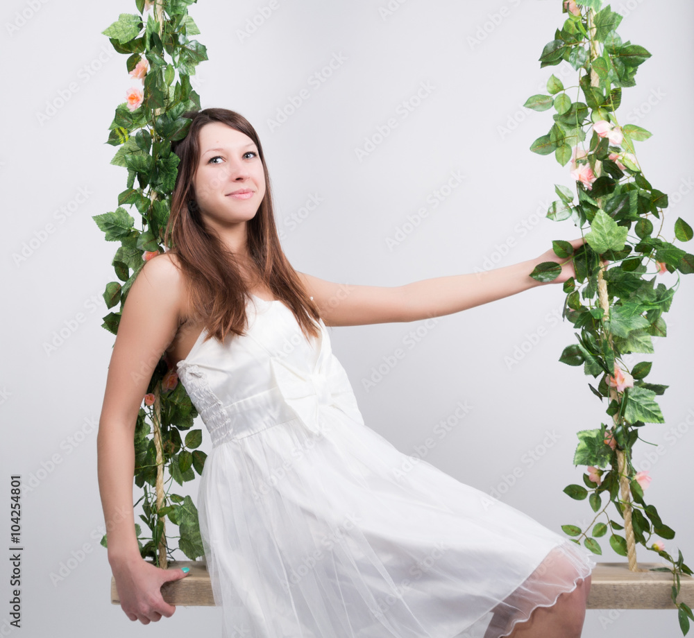 Beautiful young leggy blonde in a little white dress on a swing, wooden swing suspended from a rope hemp, rope wrapped vine and ivy Beautiful young leggy blonde in a little white dress on a swing, wooden swing suspended from a rope hemp, rope wrapped vine and ivy