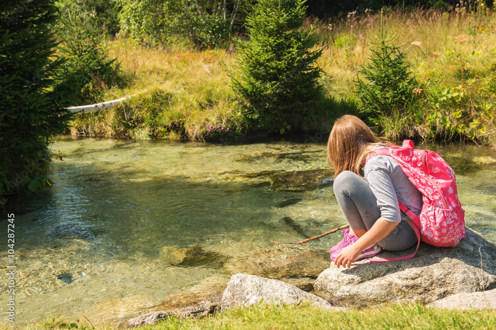 Cute little girl of 7-8 years old hiking in swiss Alps, resting by the river, wearing sport clothes, trainers and backpack, back view